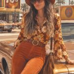 A woman in 70s style floral blouse and orange corduroy pants leaning on a vintage car.