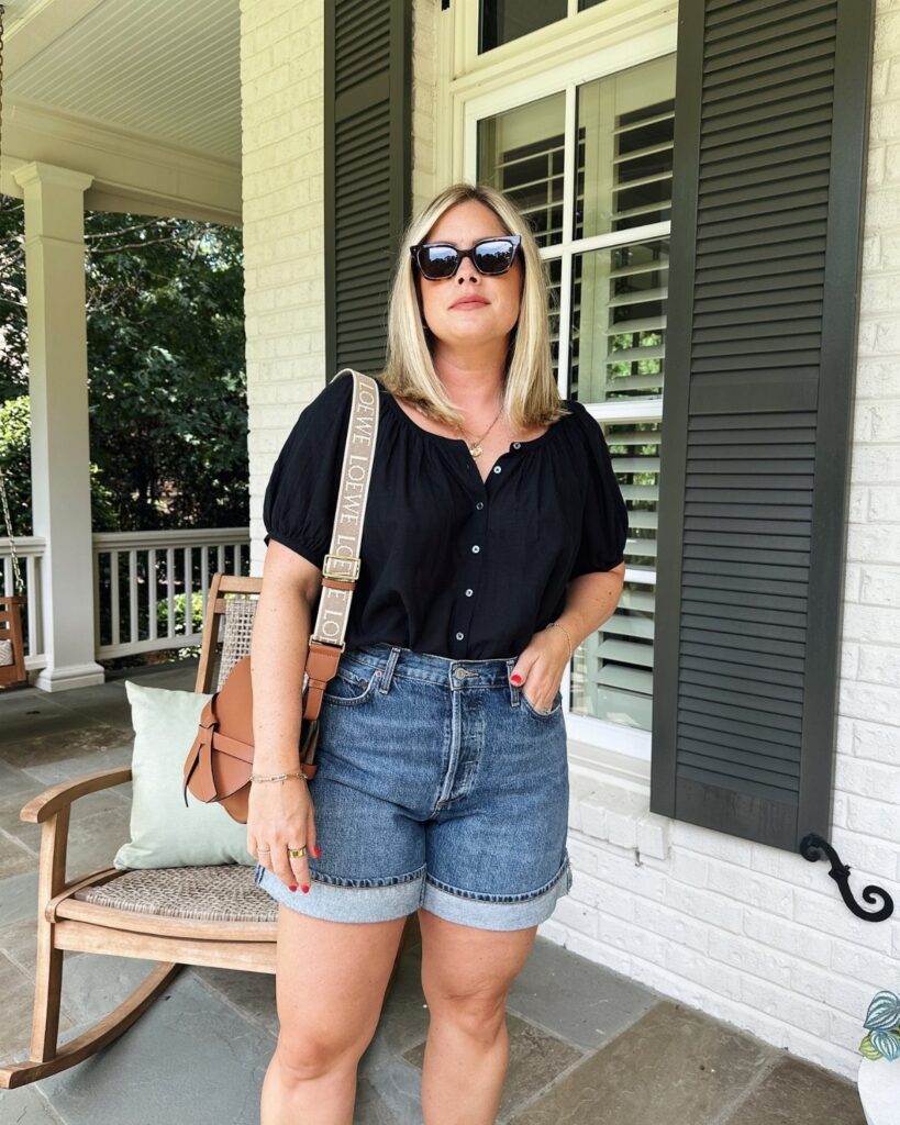 A blonde woman in a black blouse and denim shorts standing on a porch.