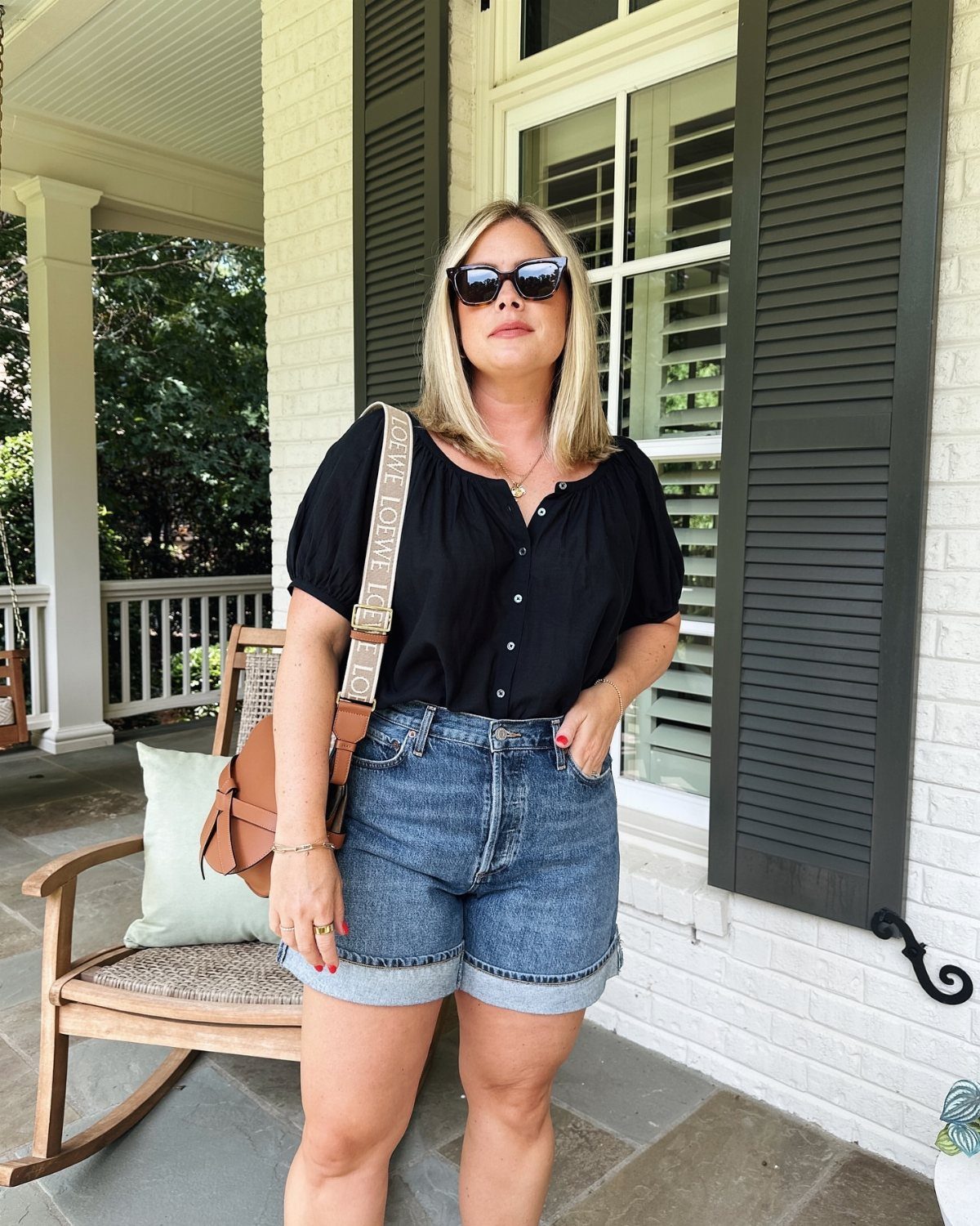 A blonde woman in a black blouse and denim shorts standing on a porch.