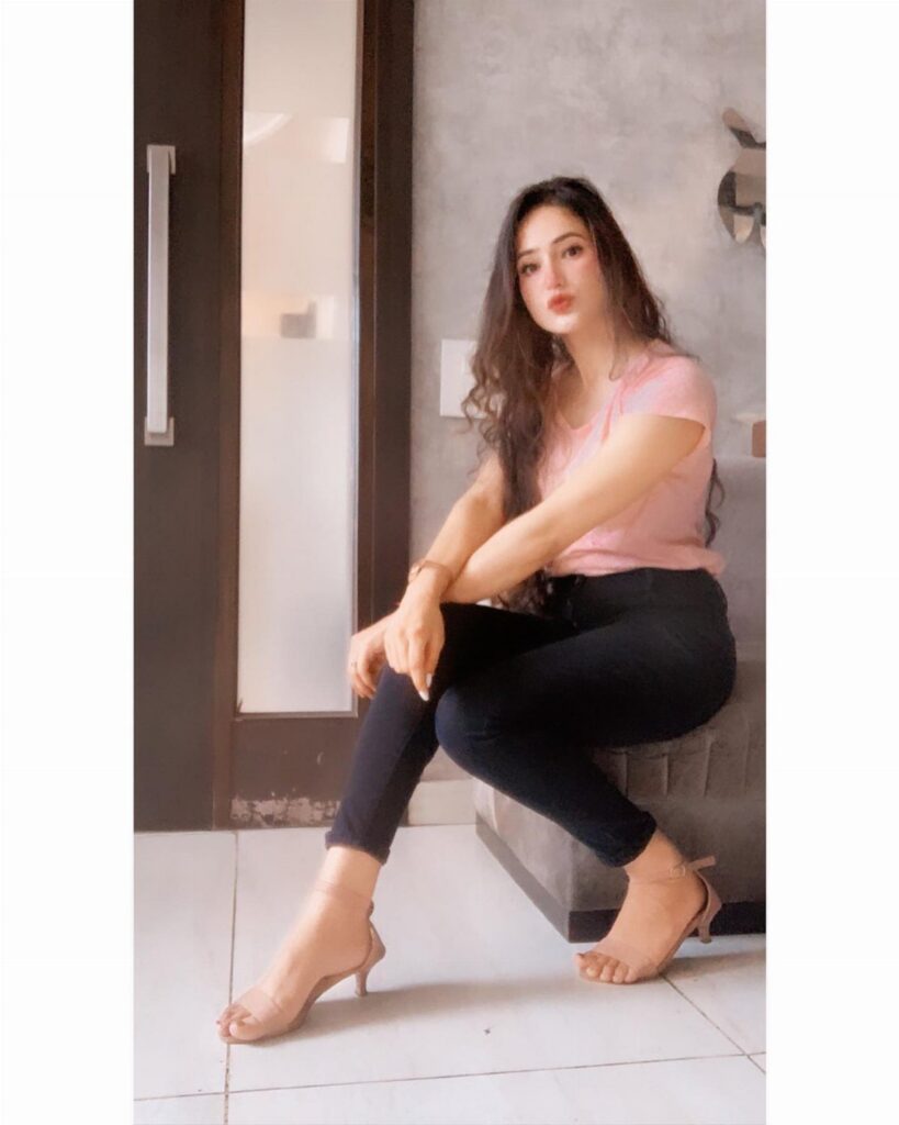 A woman sitting in a pink top and black jeans wearing nude sandals.