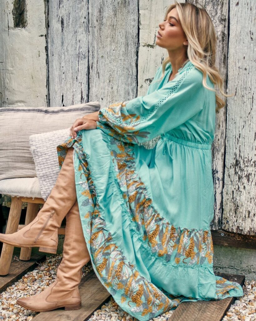 Blonde woman sitting in a turquoise floral embroidered maxi dress and tan boots.