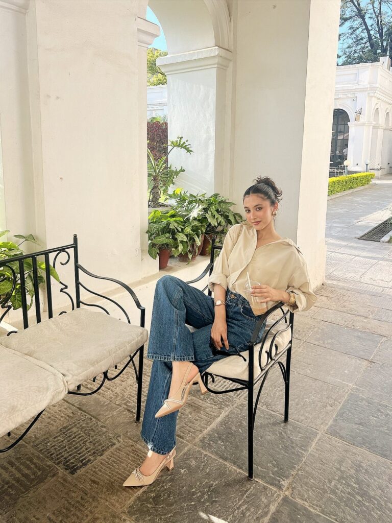 A woman wearing a beige shirt and blue jeans sitting on a bench outdoors.