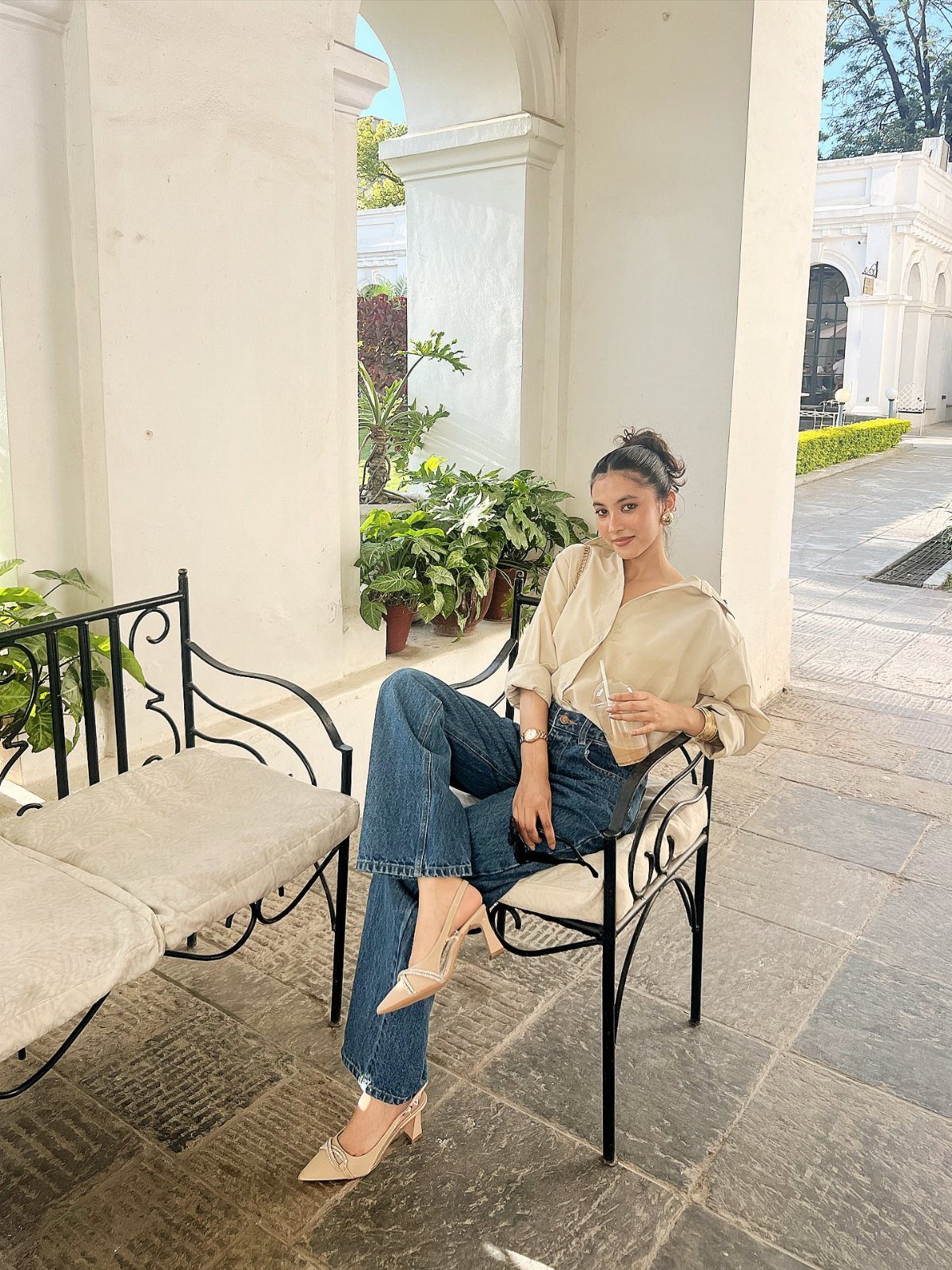 A woman wearing a beige shirt and blue jeans sitting on a bench outdoors.