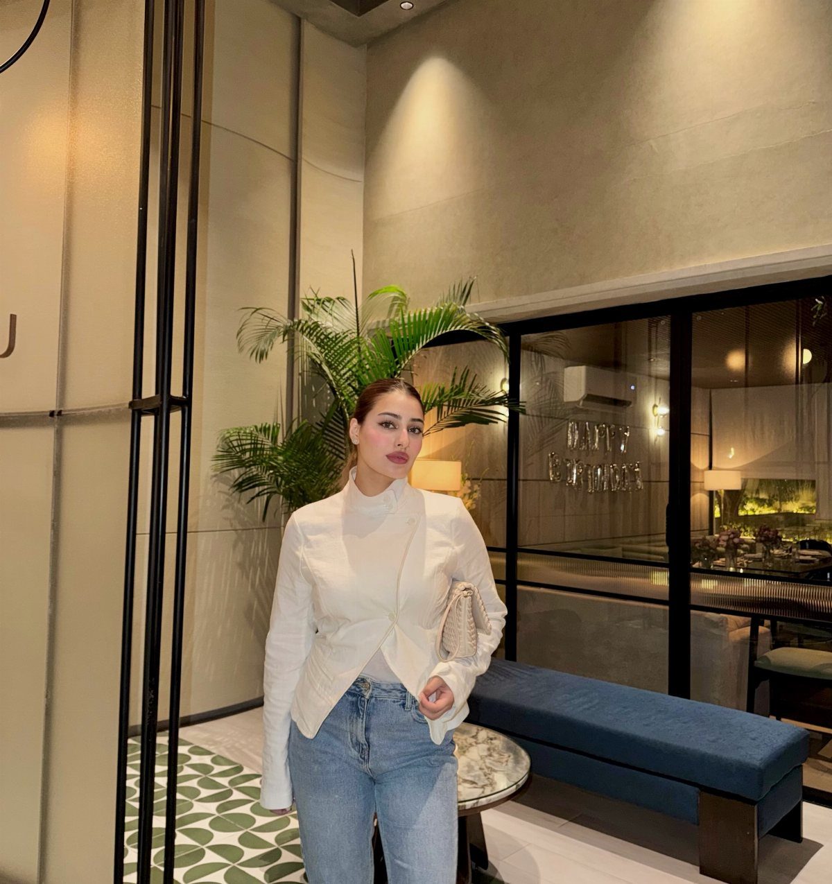 Woman in a white high-neck top and blue jeans posing in a modern interior.