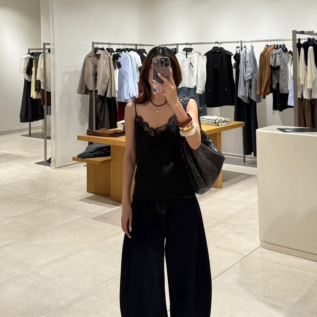 Woman in a black satin camisole and wide-leg denim trousers taking a mirror selfie.