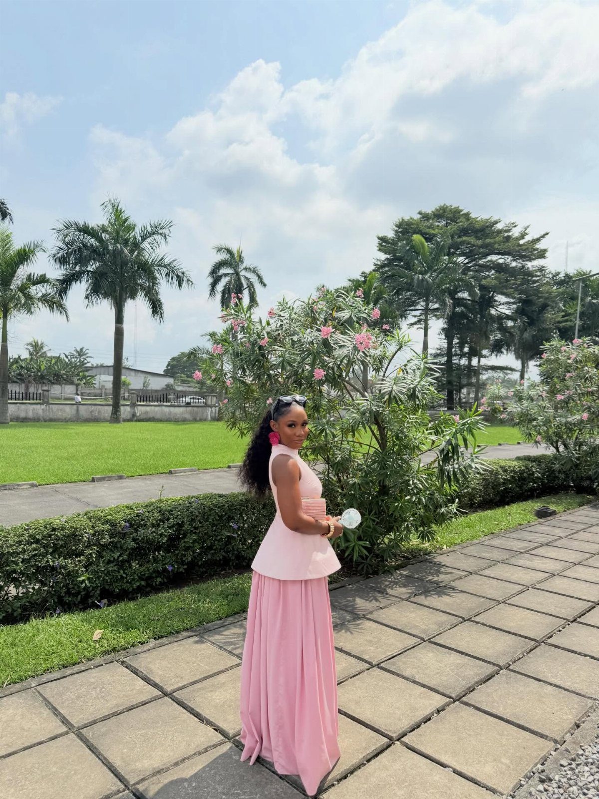 A woman in a pink peplum top and long skirt standing in a sunny park.