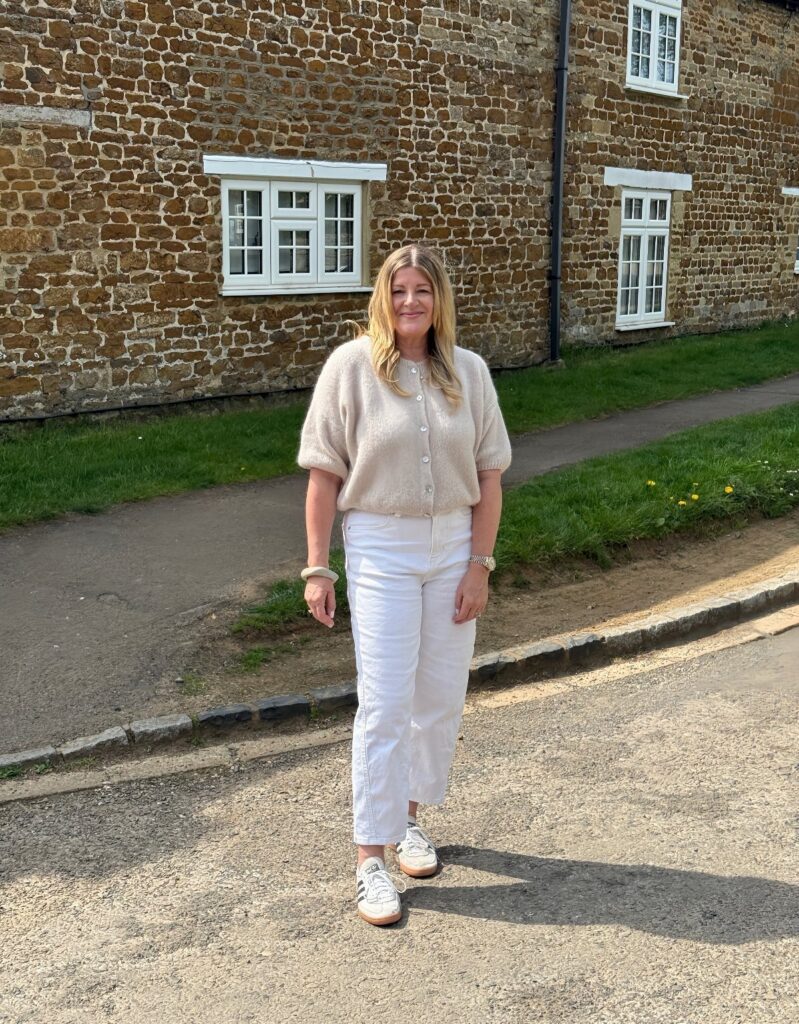 A woman wearing a cream short sleeve sweater and white trousers standing outdoors.