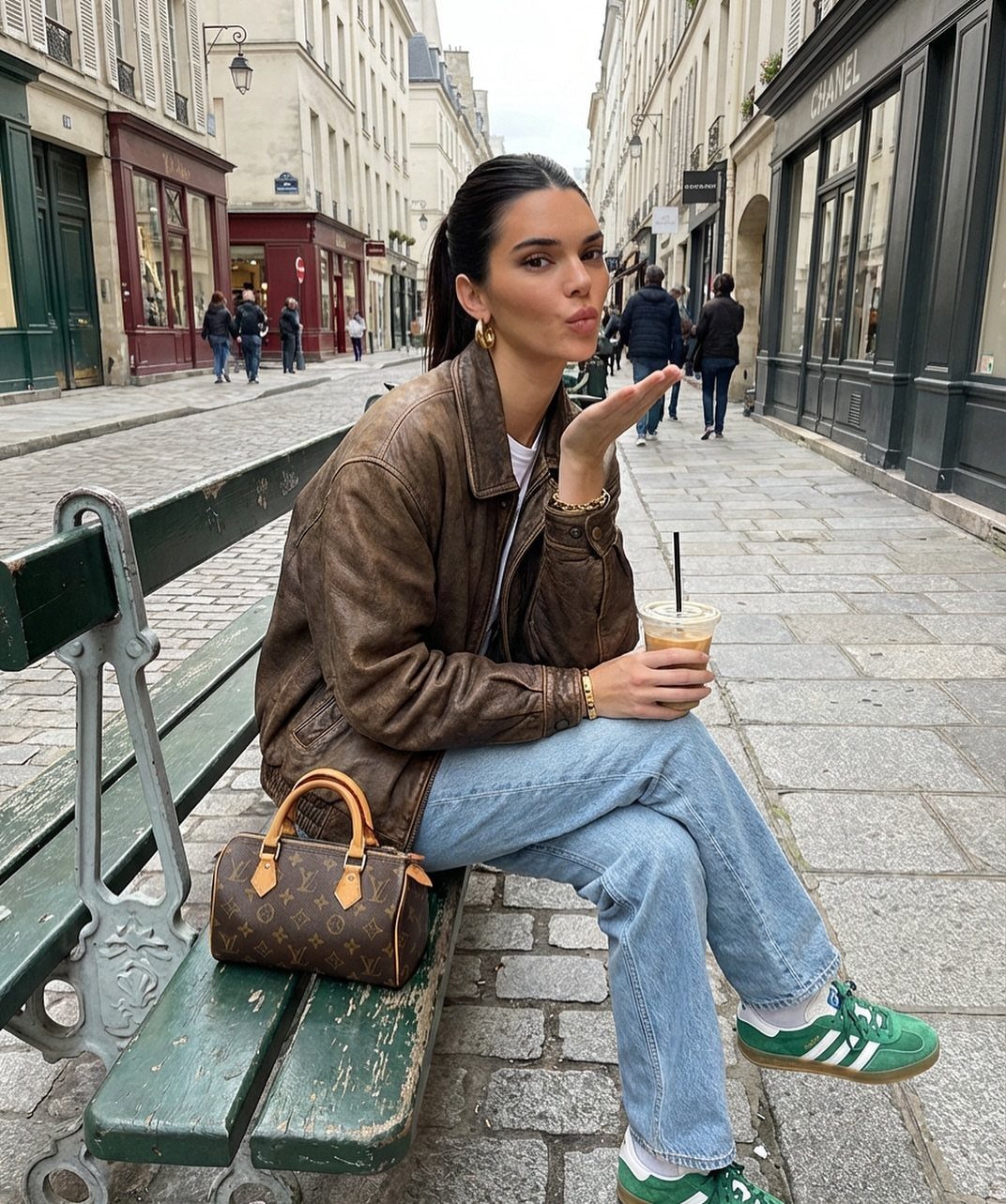 Woman in a leather jacket and blue jeans sitting on a street bench holding iced coffee.