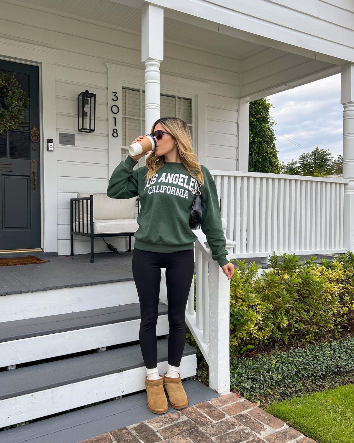 Woman in a green pullover sweatshirt, black leggings, and tan boots standing on a porch.