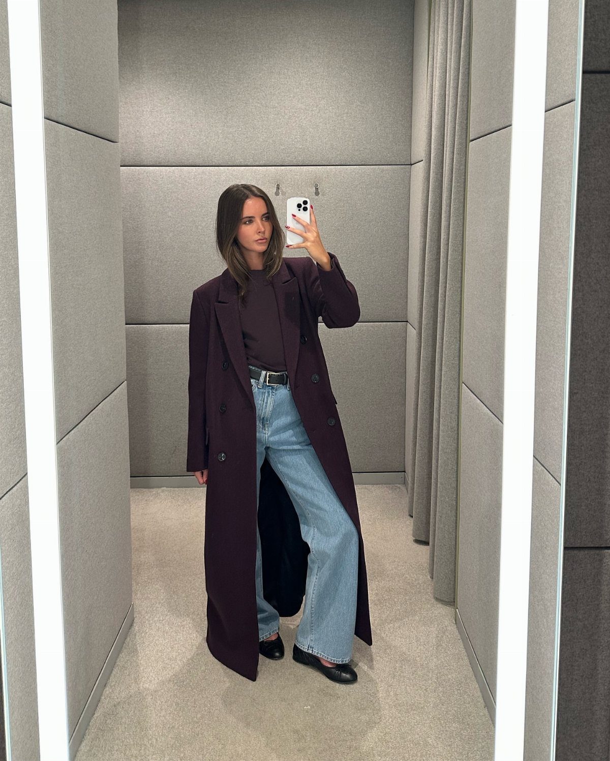 Woman in a long dark purple coat and light blue jeans taking a mirror selfie.