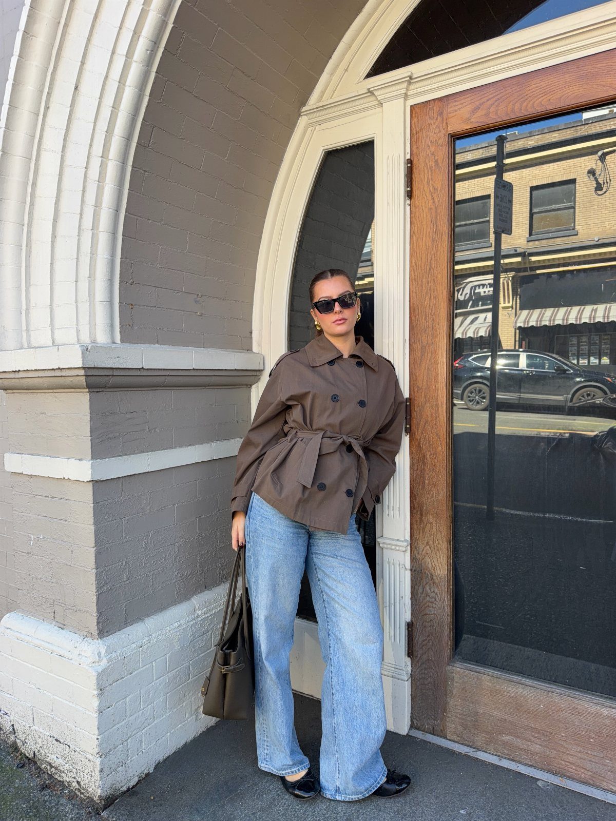 Woman in a brown belted cropped trench coat and wide-leg blue jeans standing by a doorway.
