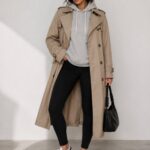 Woman wearing a long beige trench coat, grey hoodie, black leggings, and white sneakers.