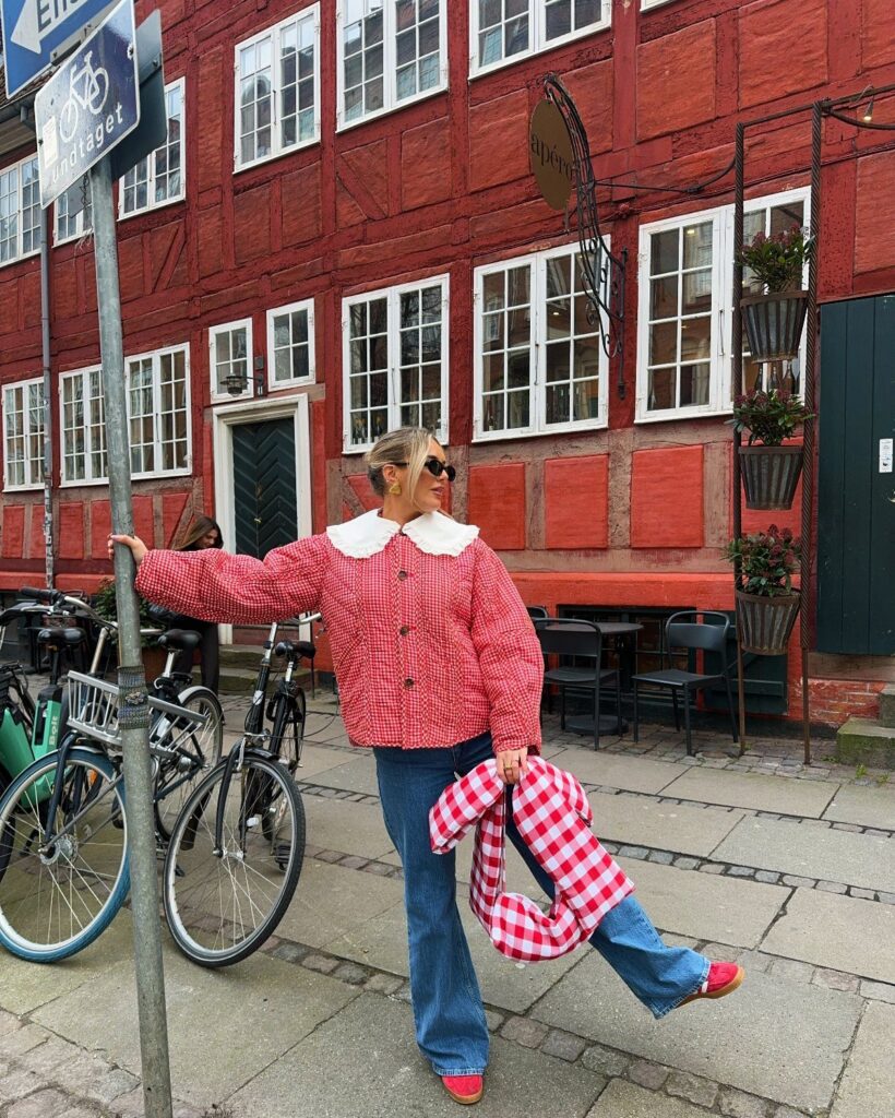 Woman in a red and white checkered jacket and blue jeans standing on a city street.