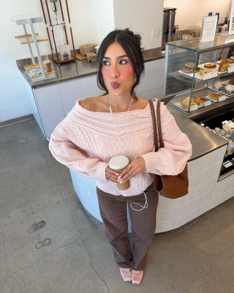 A woman in a pink knit sweater and brown pants holding a coffee cup in a cafe.