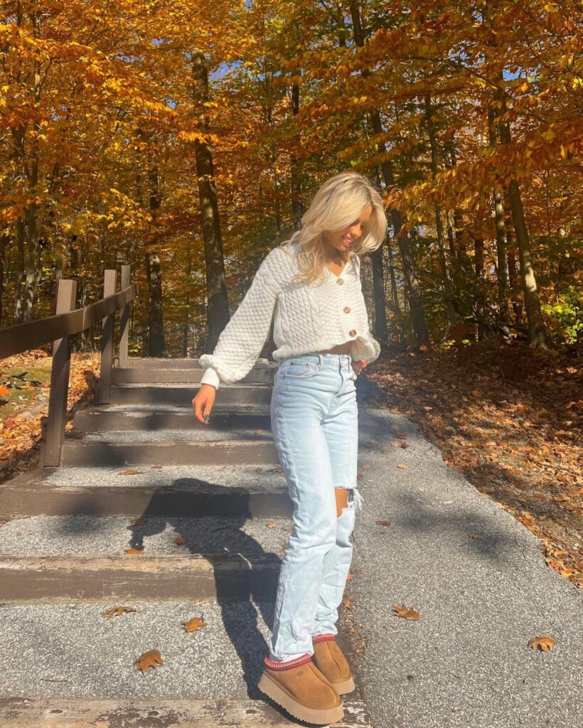 A blonde woman in a white knit sweater and jeans walking through autumn woods.