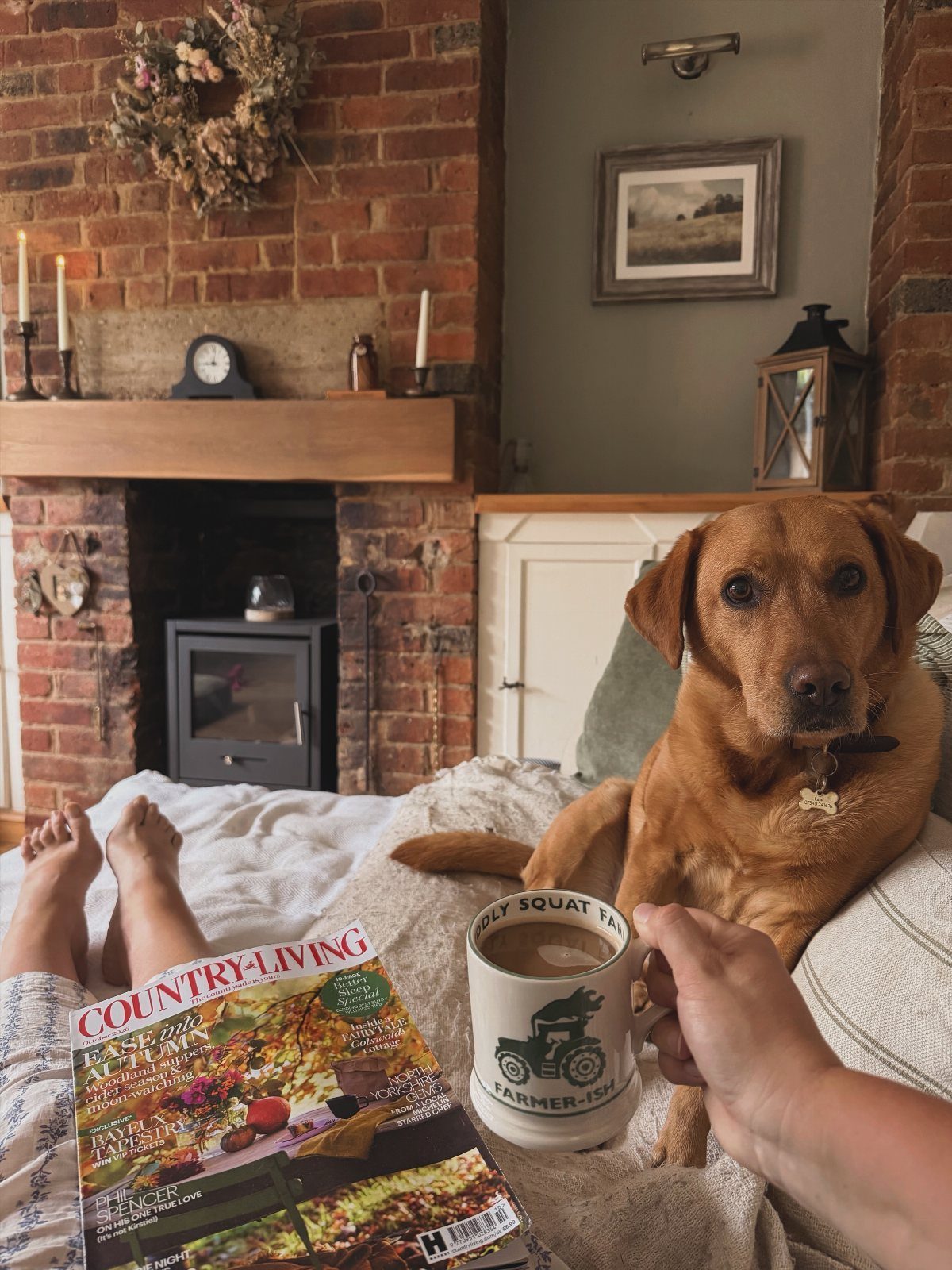A person relaxes on a bed with a dog, a magazine, and a coffee mug.