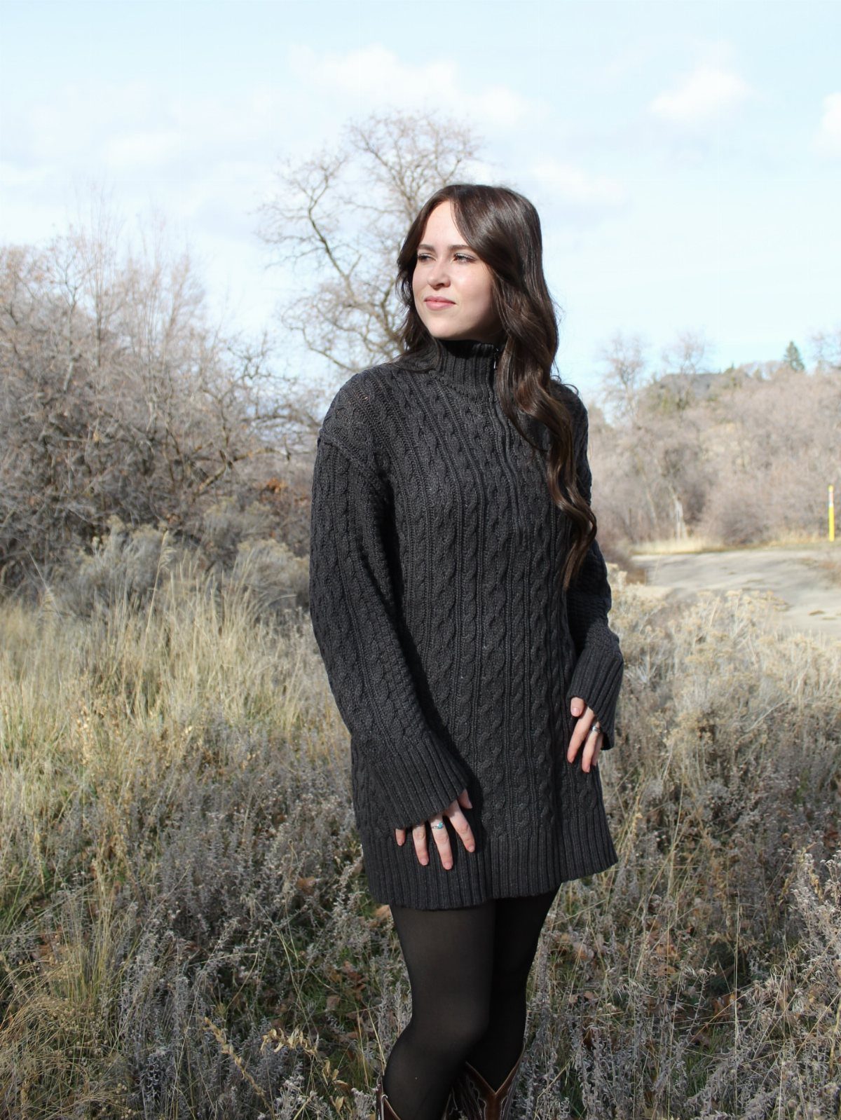 A woman wearing a dark grey cable-knit sweater dress standing in a dry grassy field.
