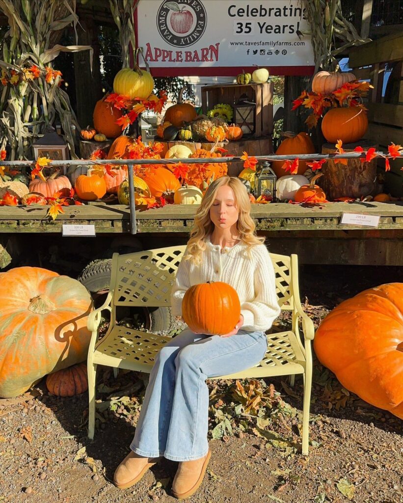 A woman wearing a white sweater and flare jeans sits on a bench holding a pumpkin.