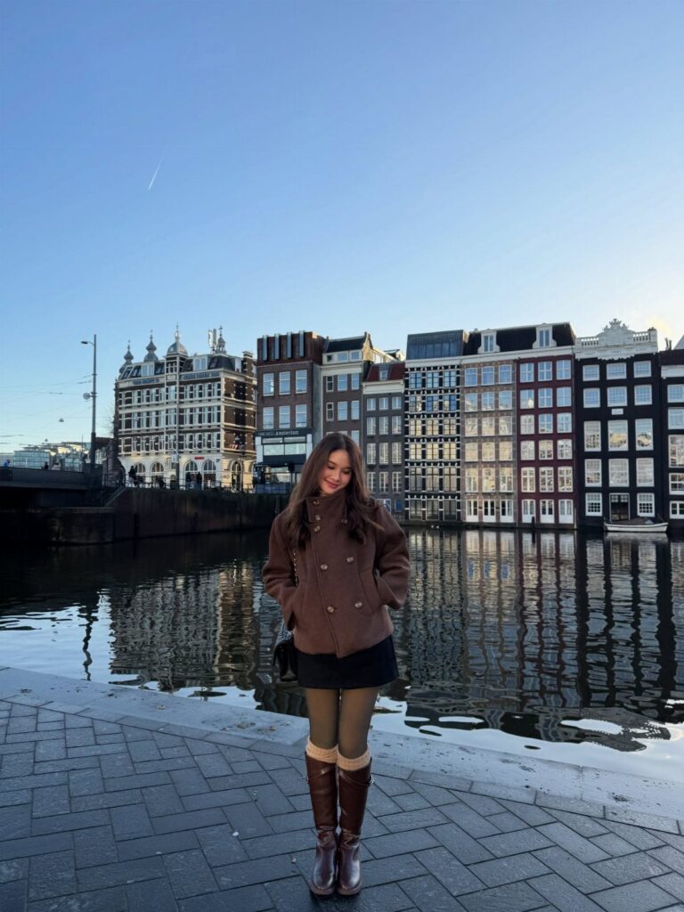 A woman wearing a brown pea coat, black skirt, green tights, and brown boots by a canal.