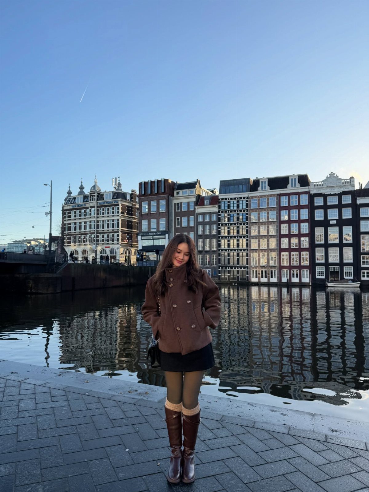 A woman wearing a brown pea coat, black skirt, green tights, and brown boots by a canal.