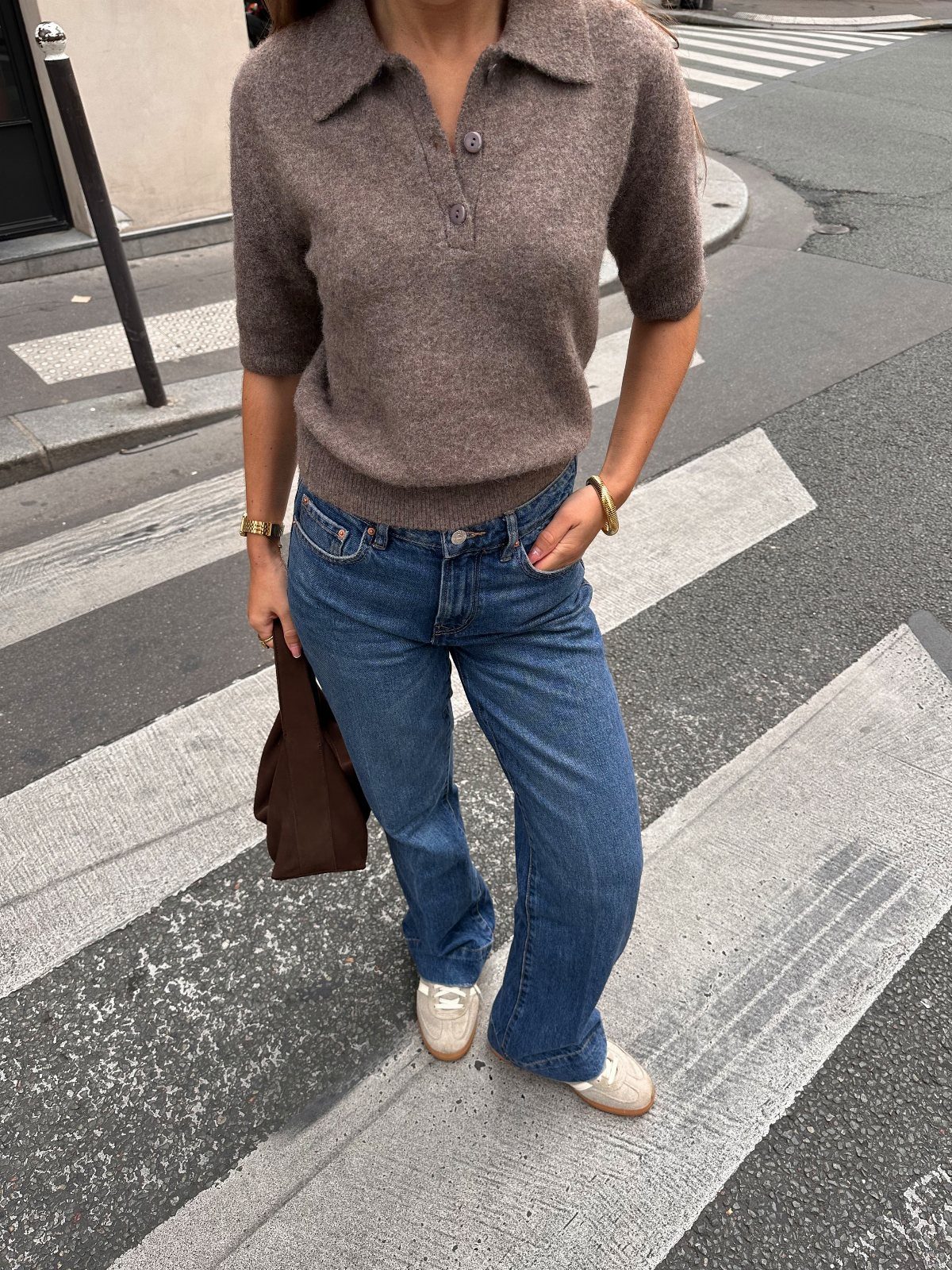 Person wearing a brown knit polo, blue jeans, and white sneakers standing on a street crosswalk.