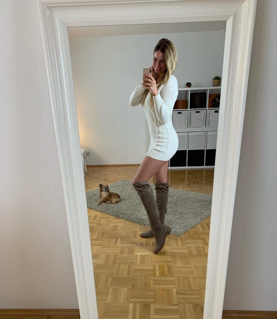 A woman posing in a white mini dress and tall taupe boots for a mirror selfie.