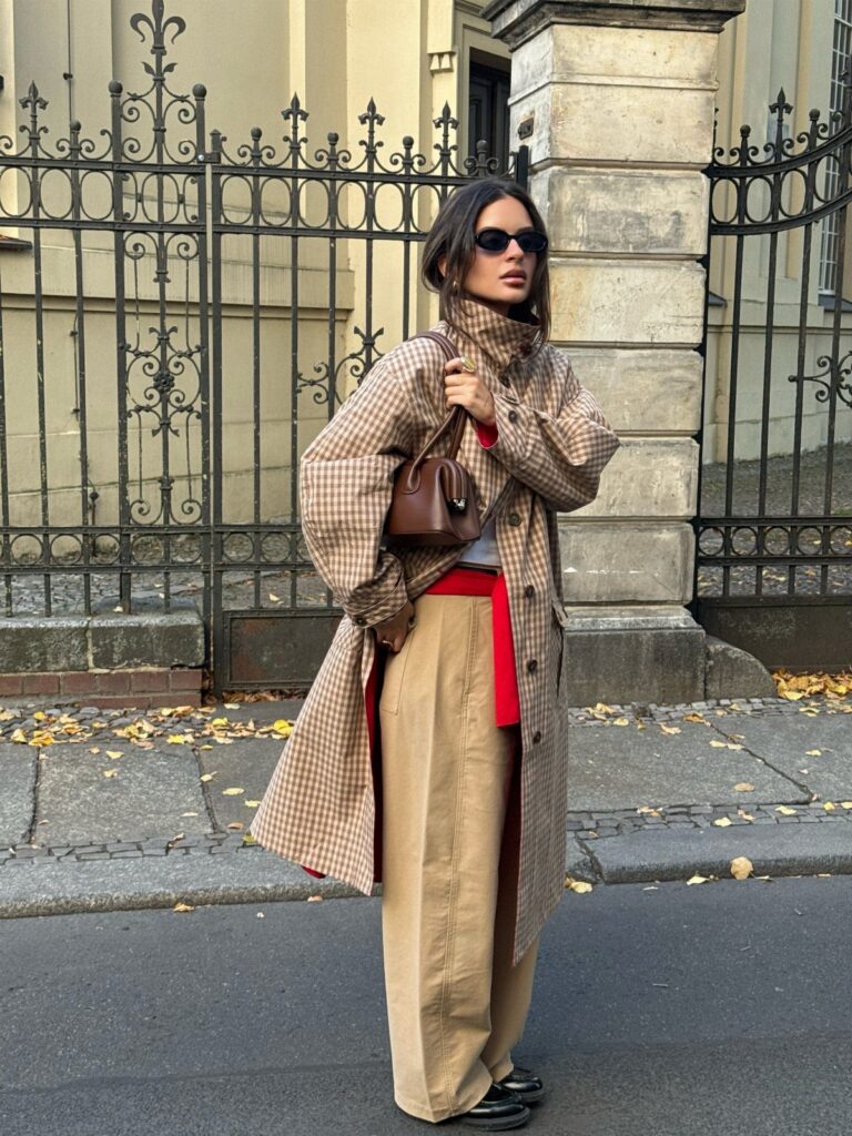 Woman wearing a long checkered trench coat, red top, and tan trousers on a city street.