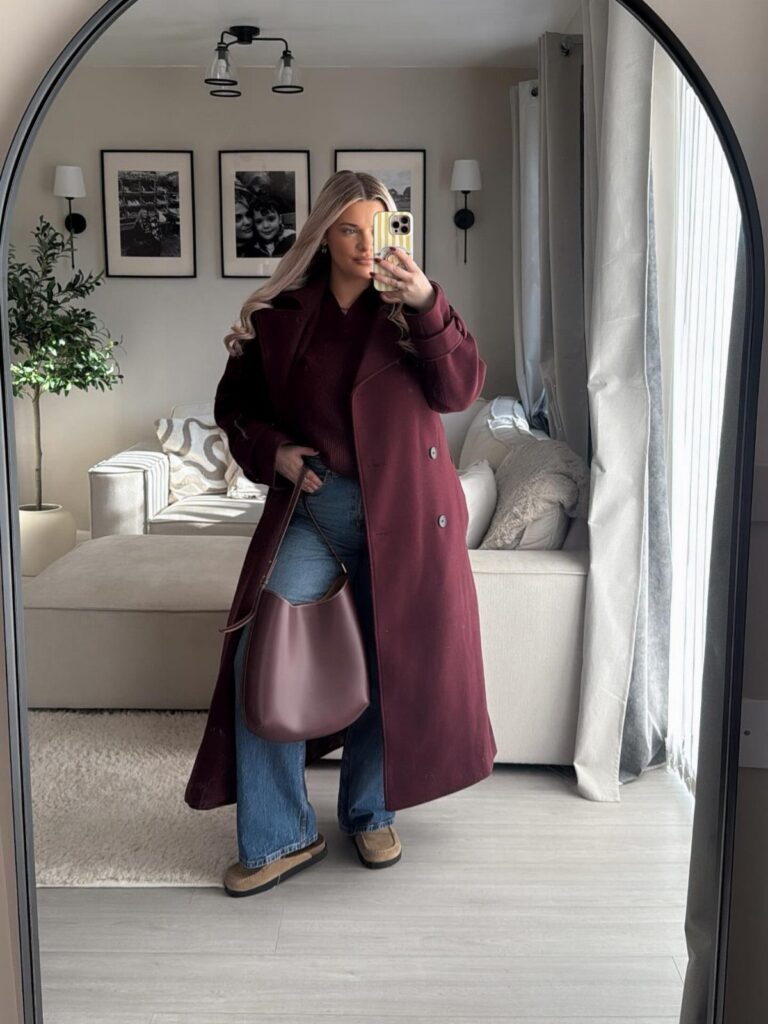 Woman in a long burgundy coat, burgundy top, and blue jeans posing for a mirror selfie.