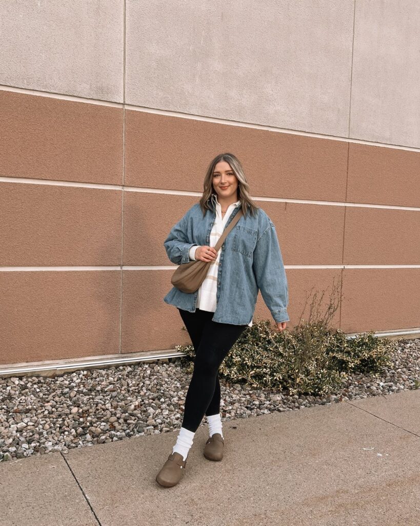 Woman wearing an oversized chambray shirt, black leggings, and brown loafers standing against a textured wall.