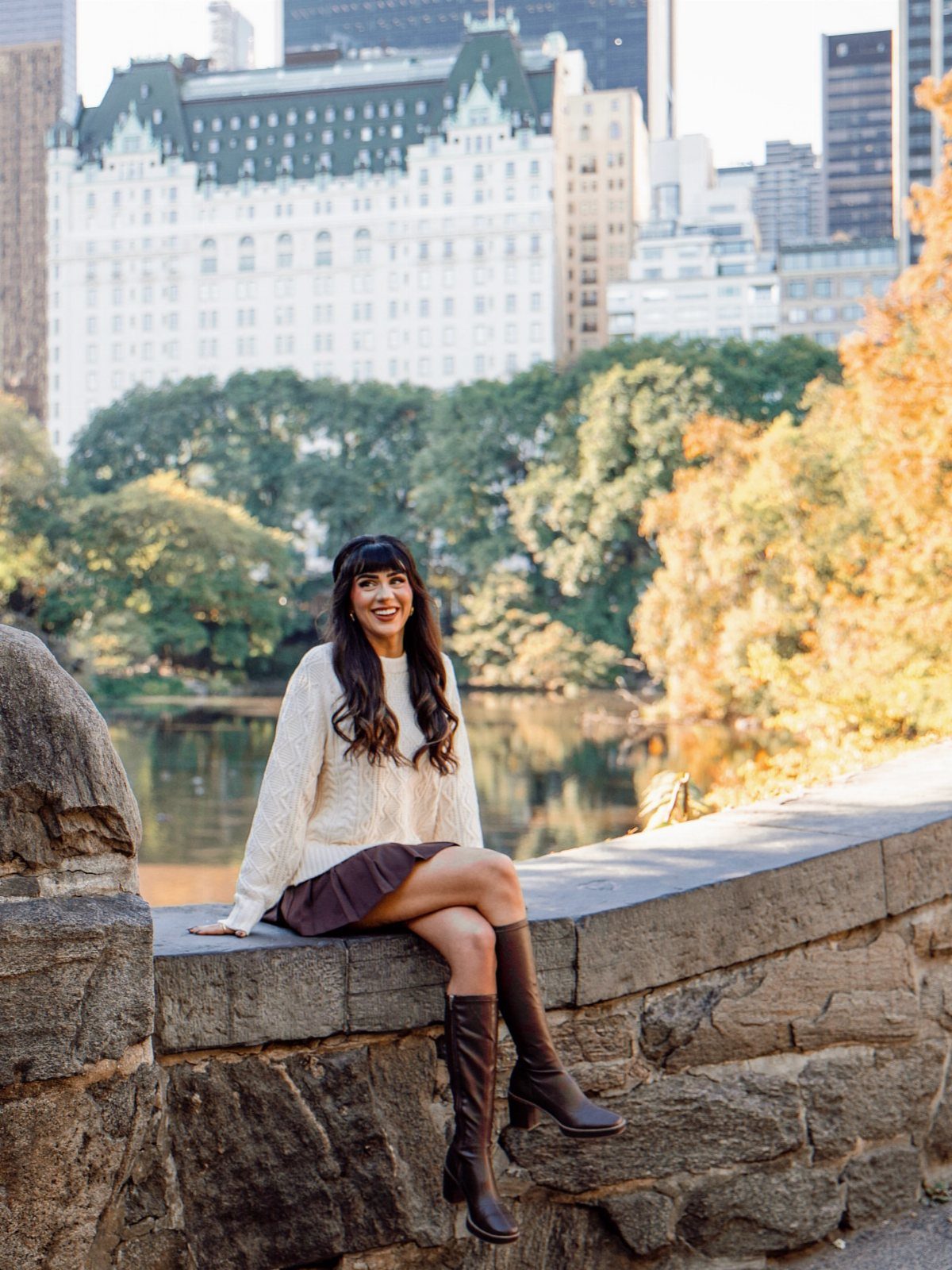 A woman wearing a cream sweater, brown skirt, and tall boots sits on a stone wall.