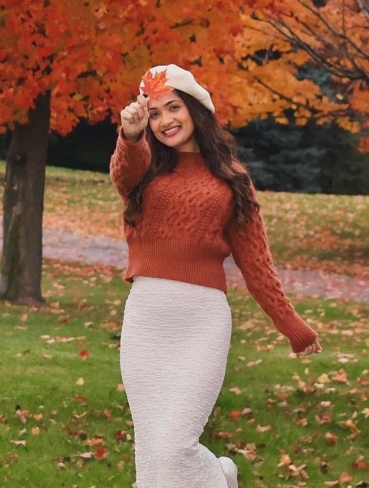 A woman in an orange sweater and cream skirt holding an autumn leaf outdoors.