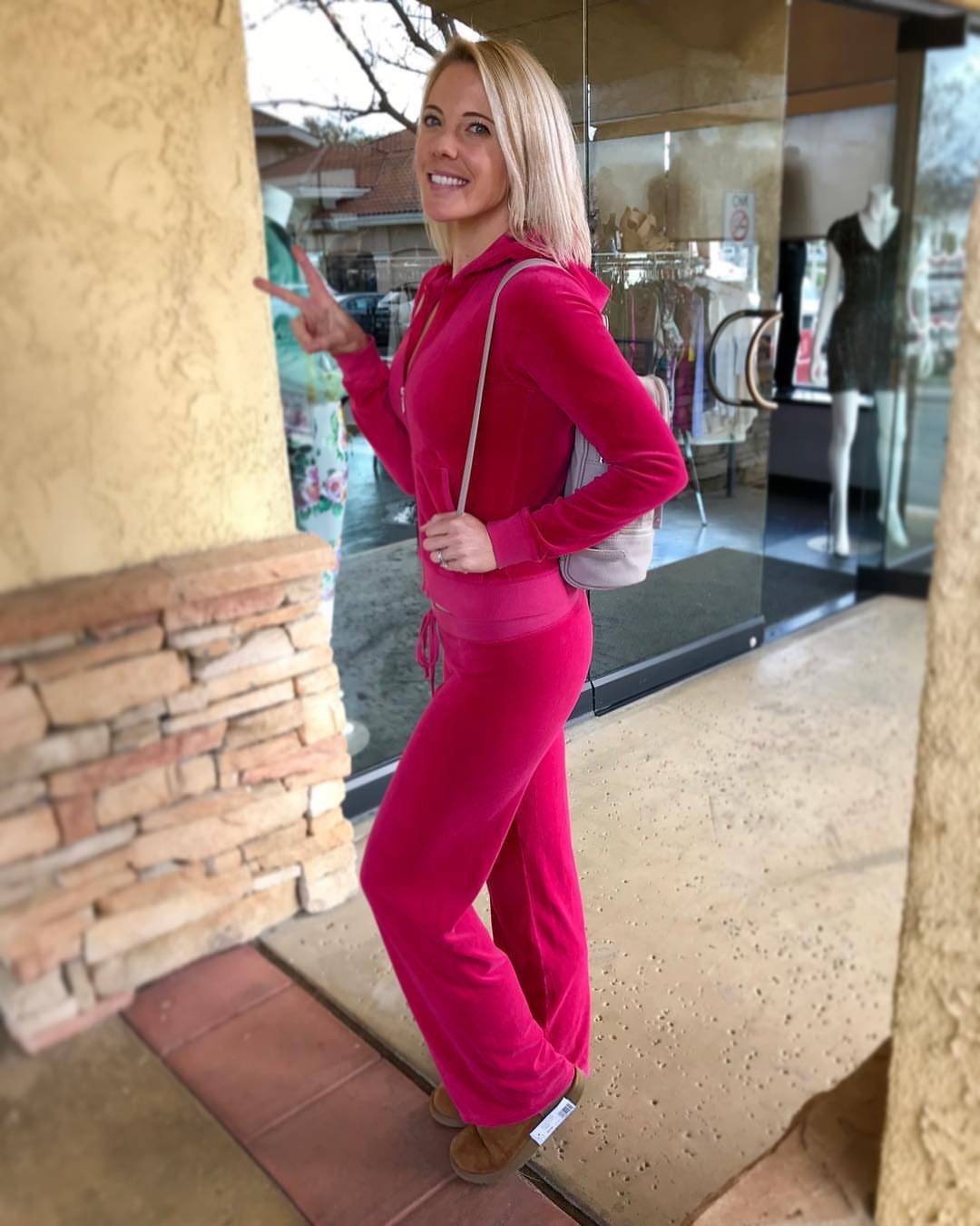 A blonde woman posing in a bright pink velour tracksuit standing near a glass storefront.