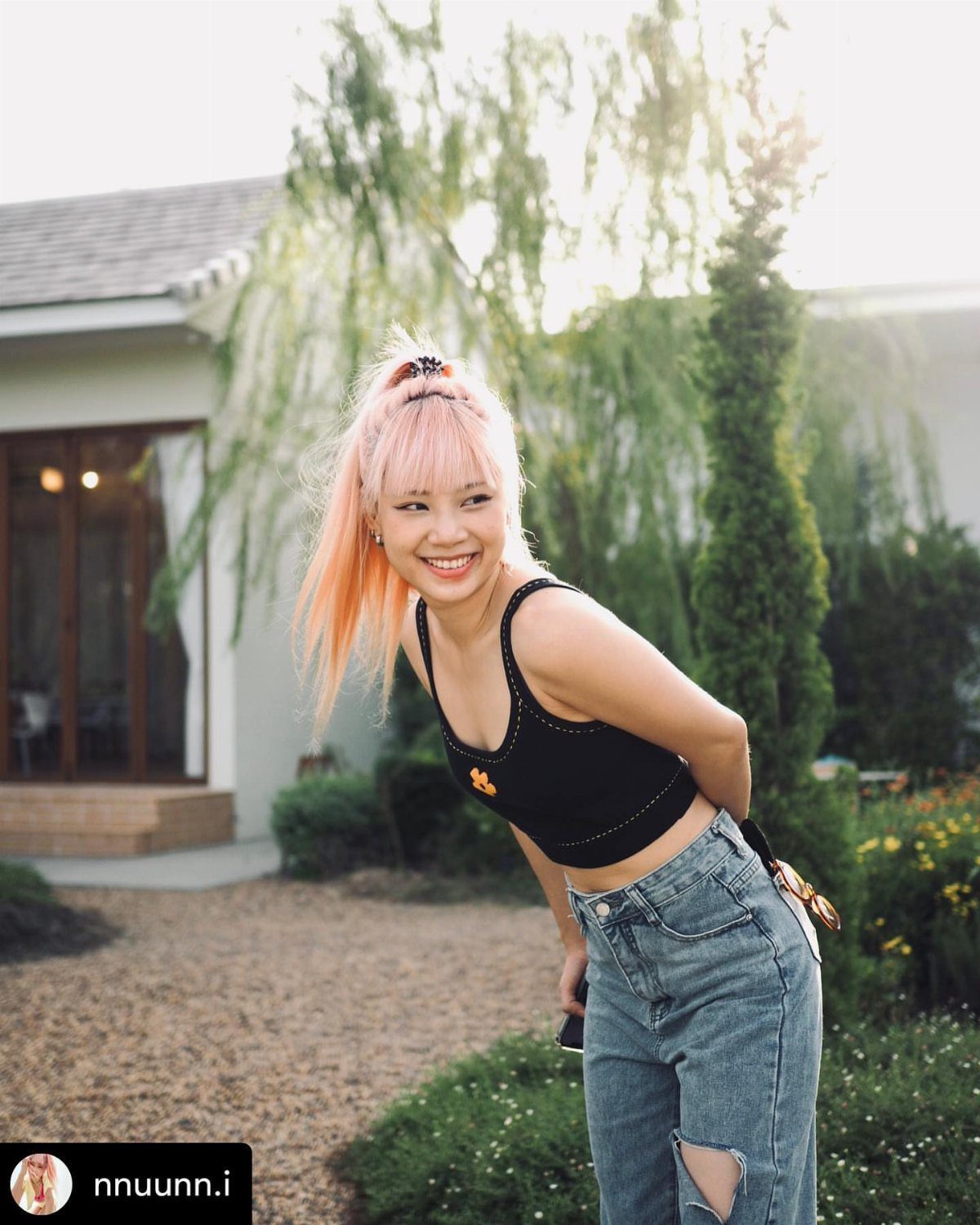 A smiling woman with pink hair wearing a black tank top and ripped blue jeans outdoors.