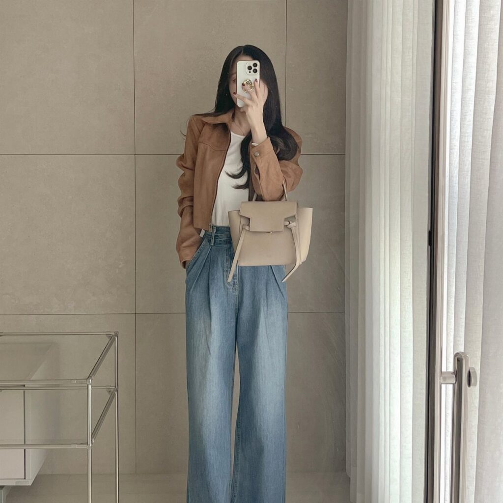 Woman in tan jacket and wide-leg blue jeans posing for a mirror selfie.