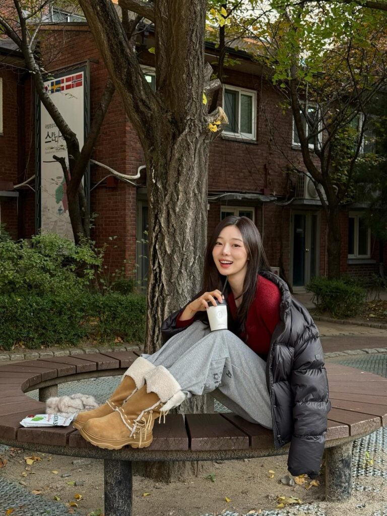 A smiling woman in a red sweater, puffer jacket, and shearling boots sitting on a bench.