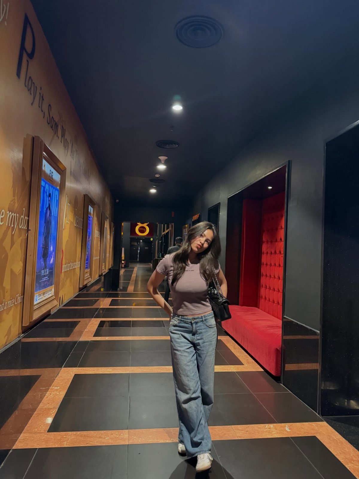 A woman in a mauve t-shirt and blue jeans posing in a cinema hallway.