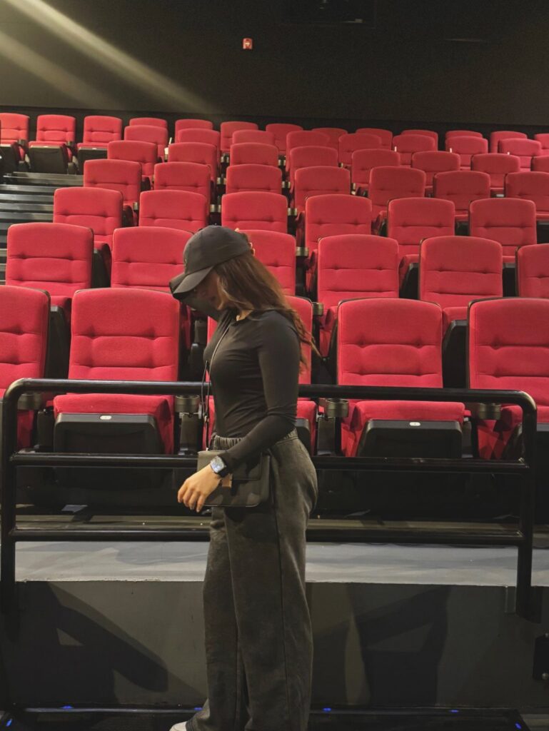 A woman in a black top and grey trousers standing in a movie theater with red seats.