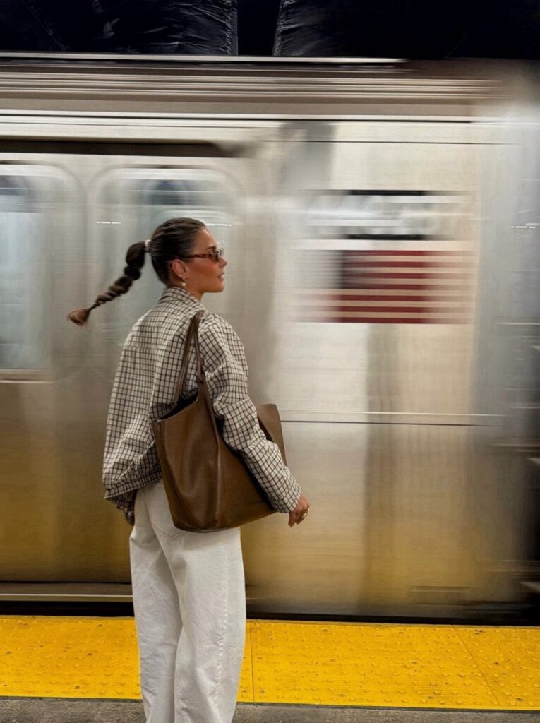 Woman in a checkered shirt and white trousers standing on a subway platform near a moving train.
