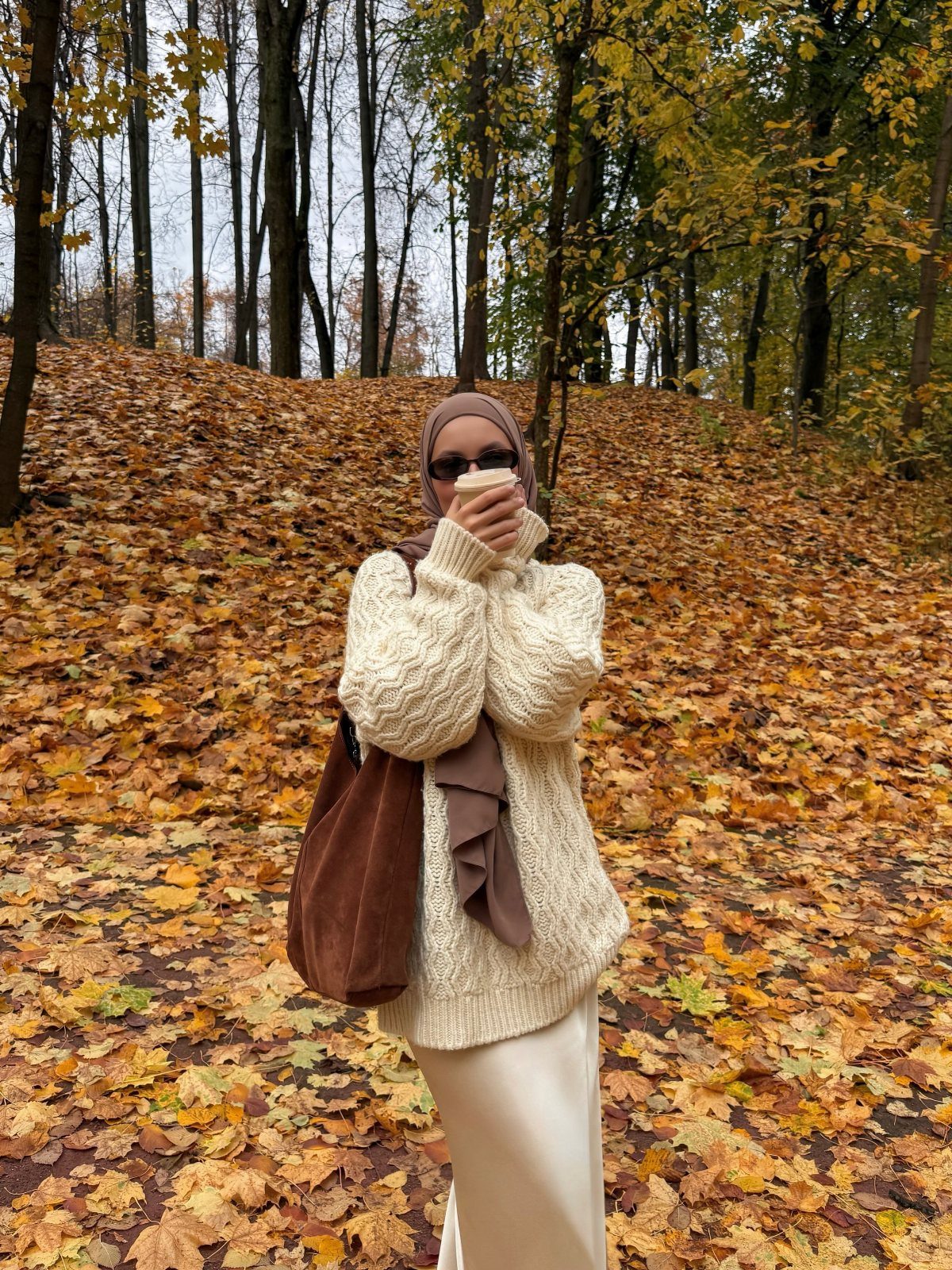 A person in a cream cable-knit sweater standing in a forest of autumn leaves.