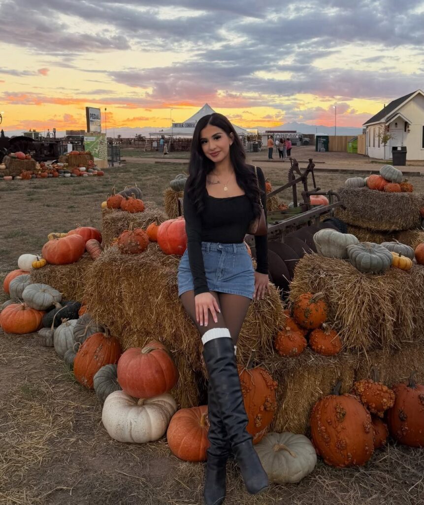 A woman in a black top and denim skirt sitting among pumpkins on hay bales.
