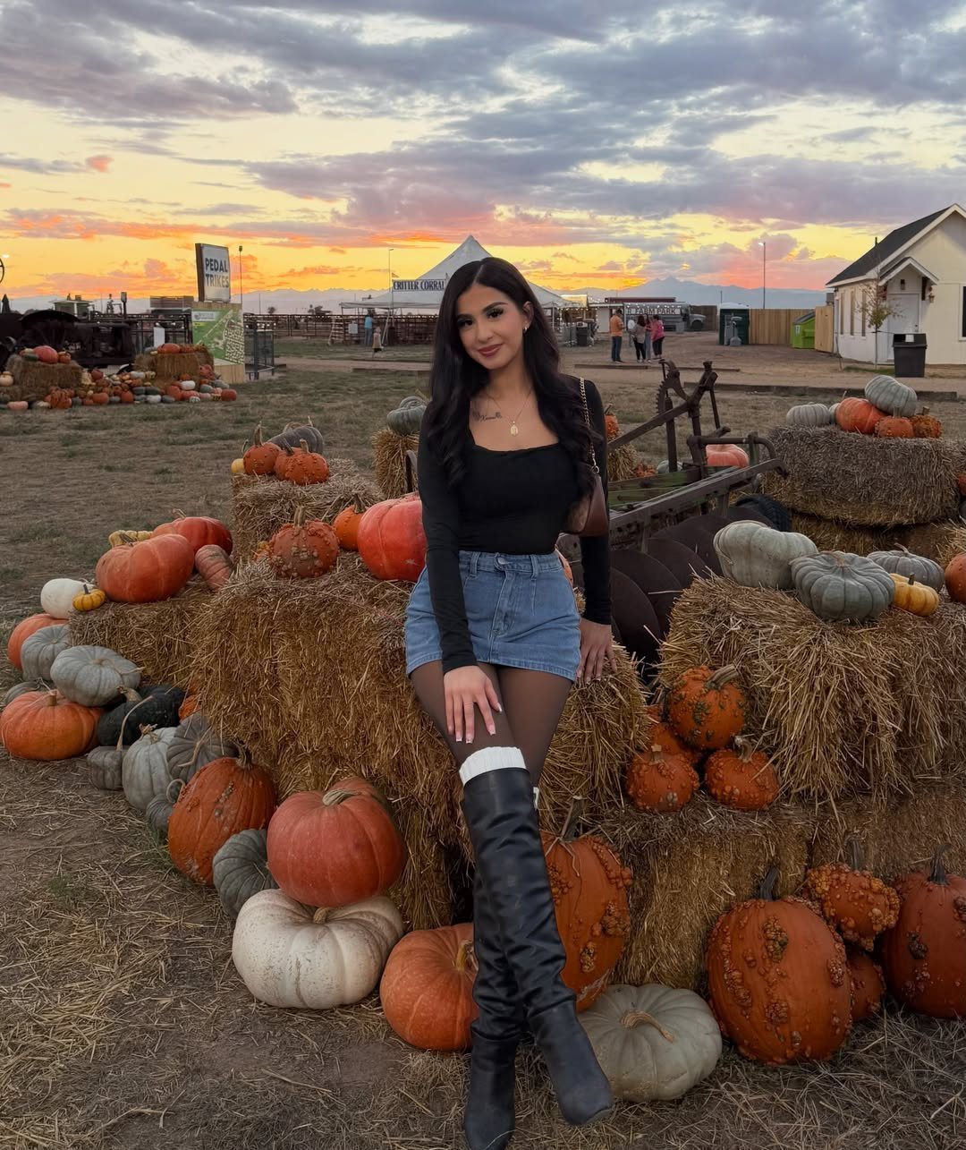 A woman in a black top and denim skirt sitting among pumpkins on hay bales.