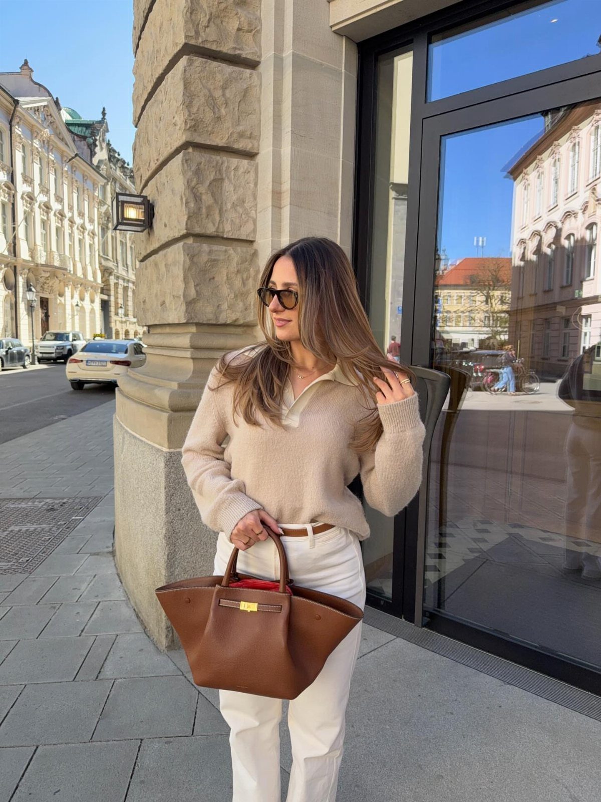 Woman in beige sweater and white trousers holding a large brown leather handbag.