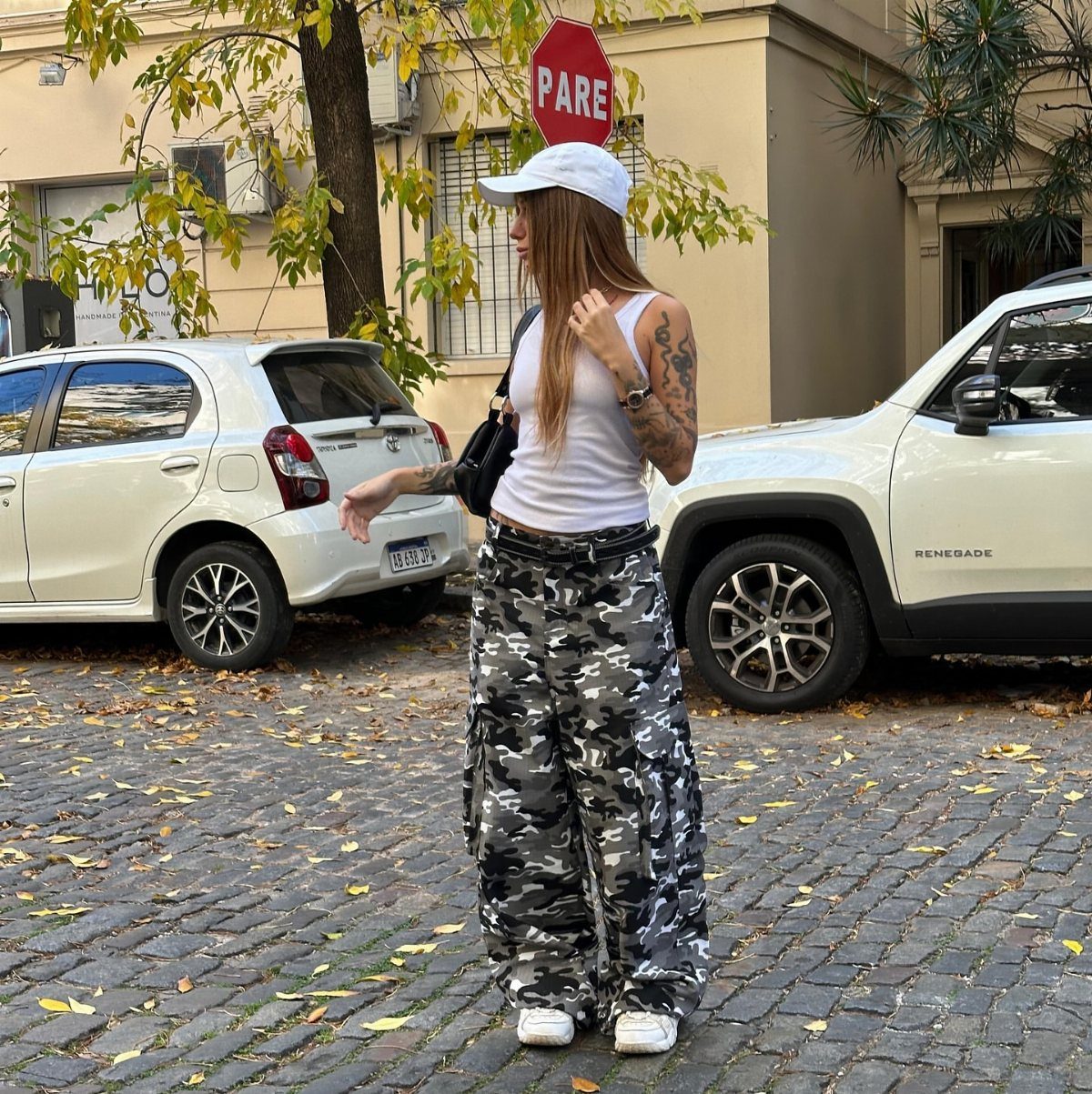 A woman wearing camouflage wide-leg cargo pants and a white tank top standing on a cobblestone street.