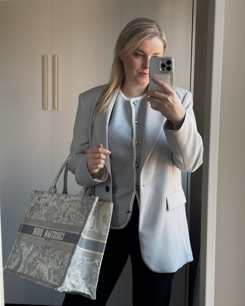 A blonde woman posing in a mirror wearing a grey blazer, white top, and black trousers.