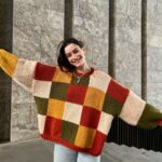 A smiling woman wearing a colorful checkered knit sweater and light blue jeans outdoors.