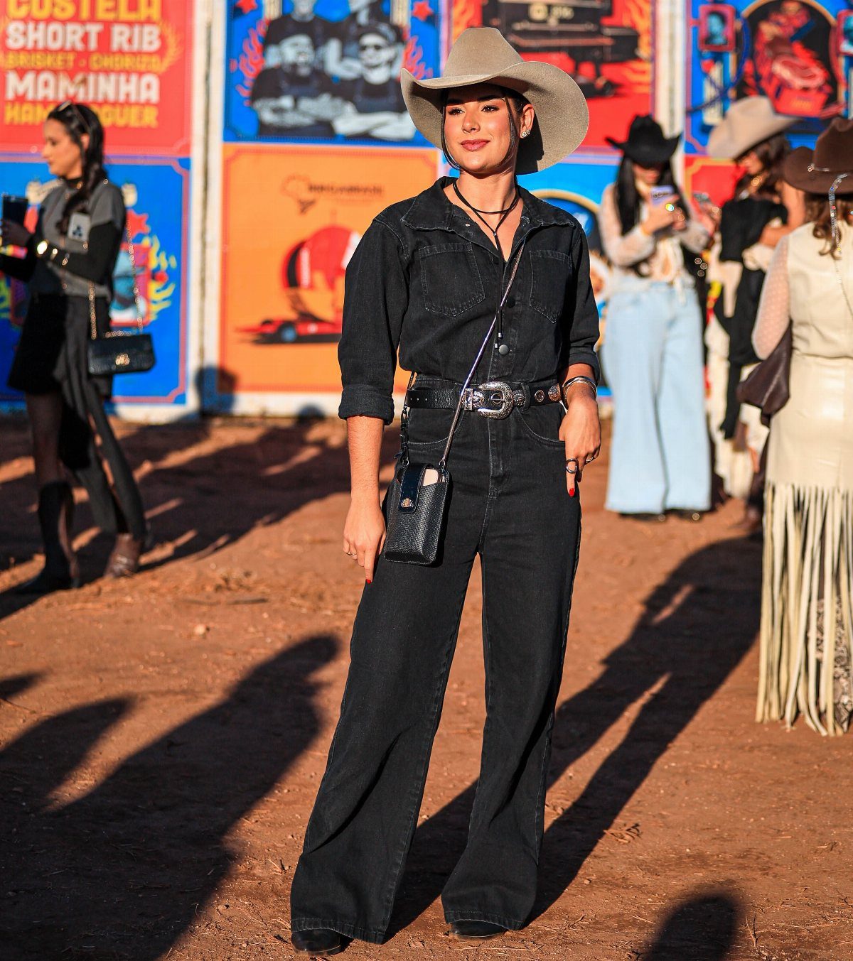 Woman in a black denim jumpsuit and cowboy hat standing outdoors against colorful posters.