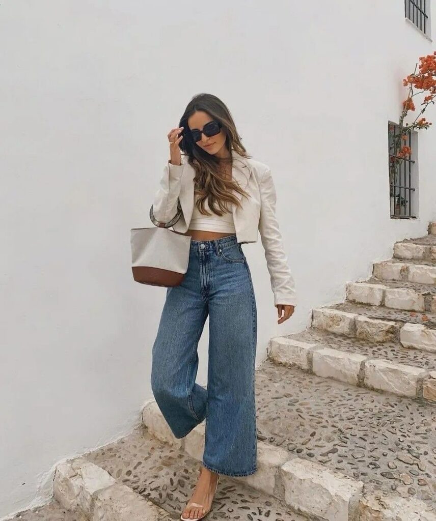 Woman wearing blue wide leg jeans, a white cropped top, and a white blazer on stone stairs.