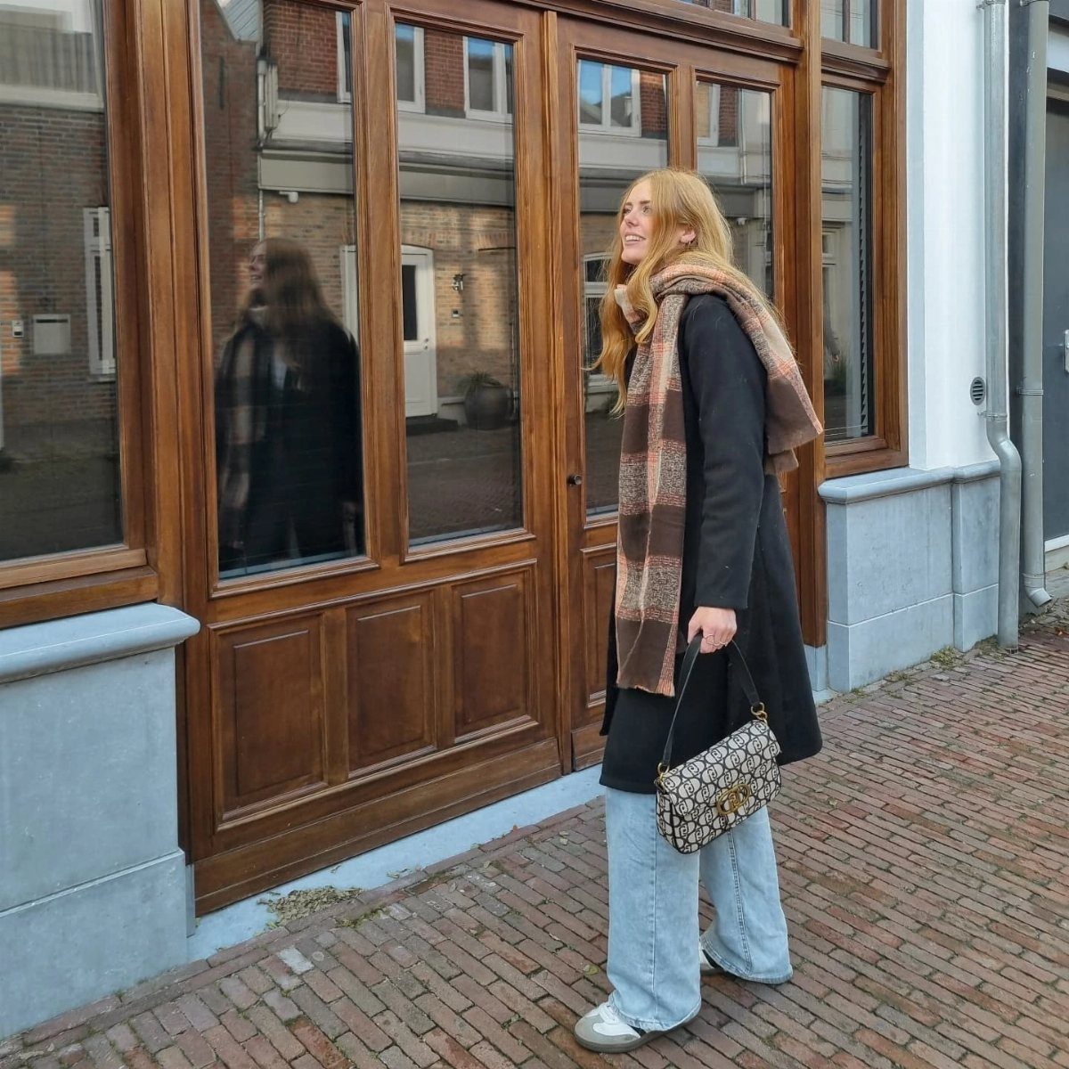 A woman walking past a wooden storefront wearing a black coat, blue jeans, and a patterned scarf.