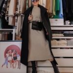 Woman wearing a beige sweater dress, black coat, and black boots standing in a bedroom.