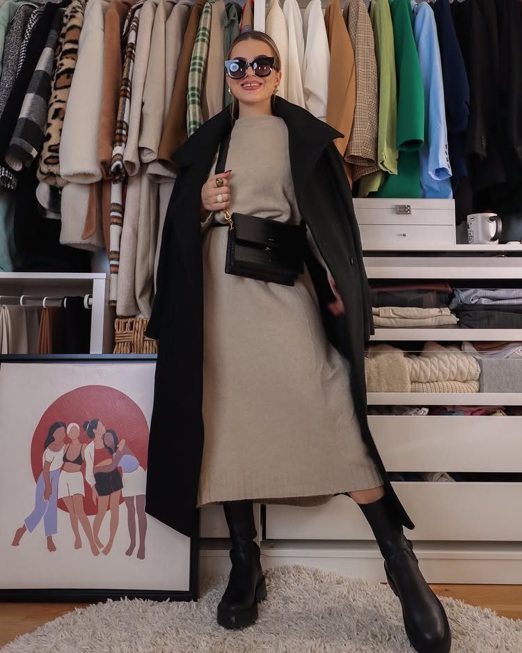 Woman wearing a beige sweater dress, black coat, and black boots standing in a bedroom.